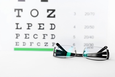 Photo of Glasses with corrective lenses on table against eye chart