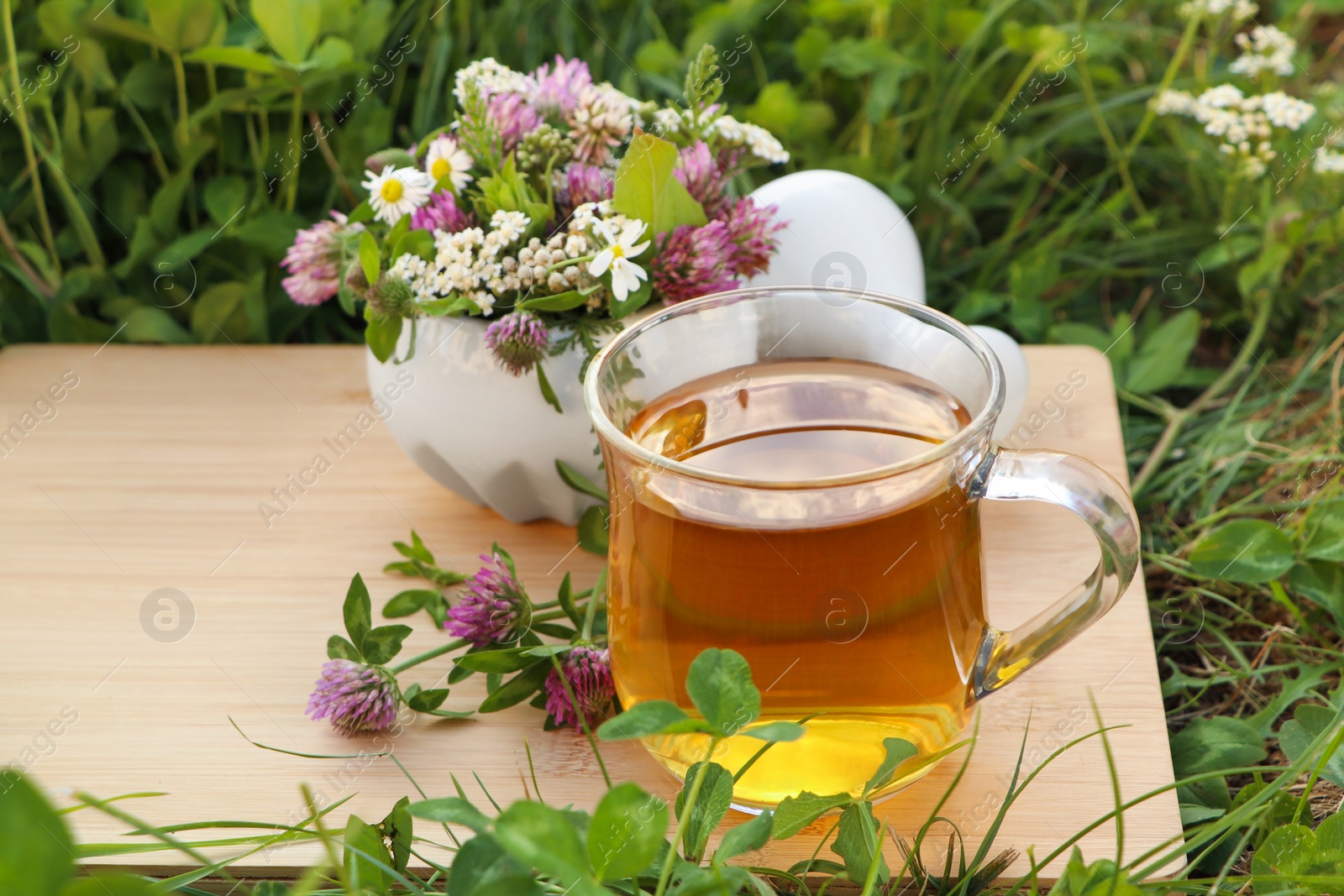 Photo of Cup of aromatic herbal tea, pestle and ceramic mortar with different wildflowers on green grass outdoors