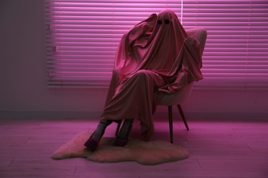 Photo of Glamorous ghost. Woman in sheet and high heel shoes on armchair in pink light