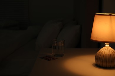 Photo of Insomnia treatment. Glass of water and pills on bedside table in bedroom at night