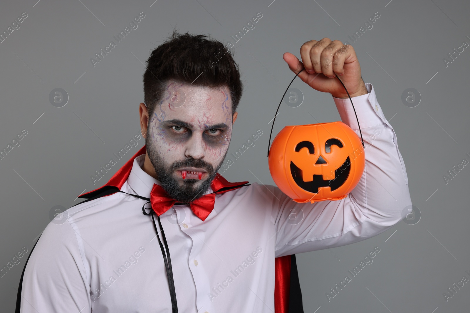 Photo of Man in scary vampire costume with fangs and pumpkin bucket on light grey background. Halloween celebration