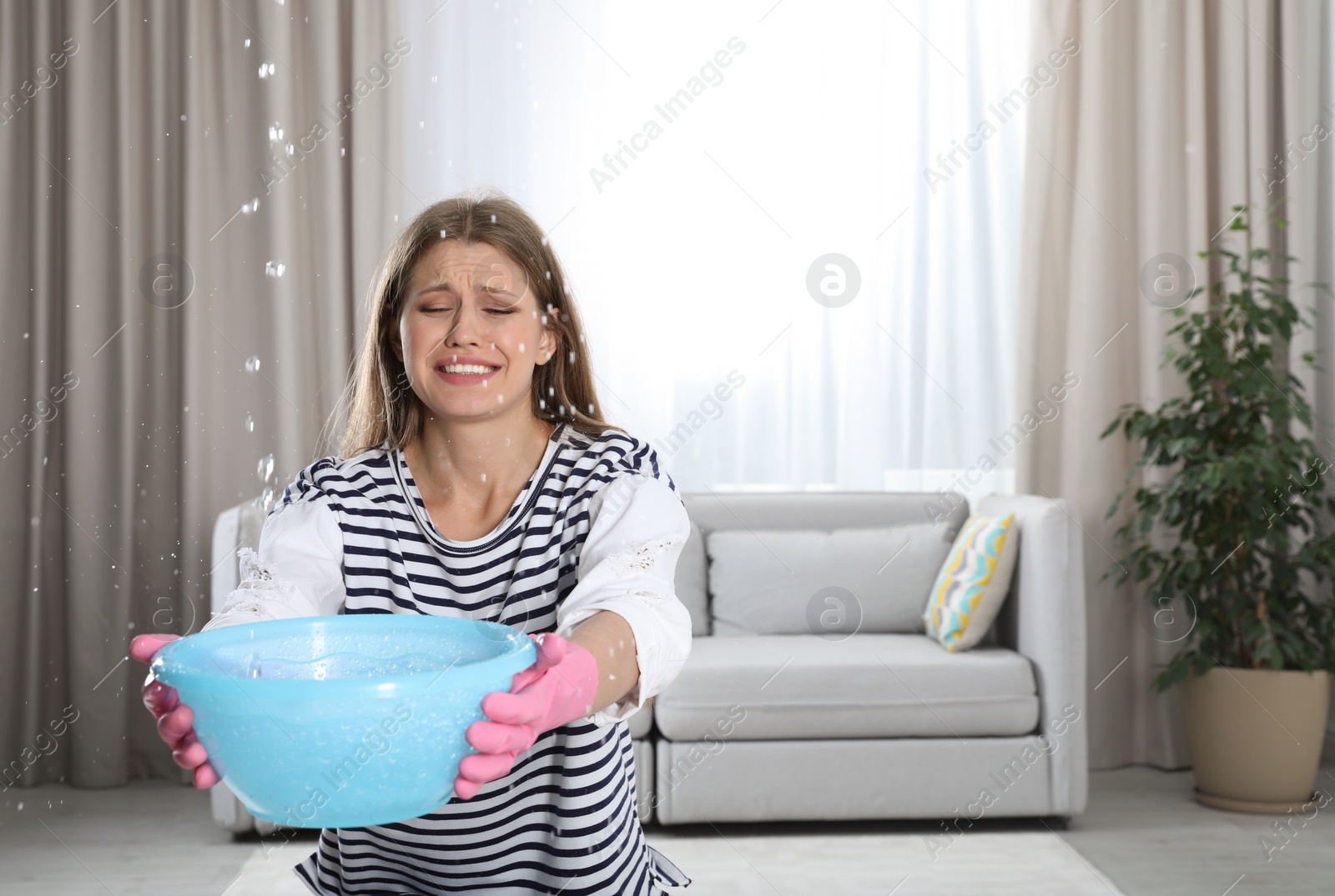 Photo of Emotional young woman collecting water leaking from ceiling in living room. Plumber service