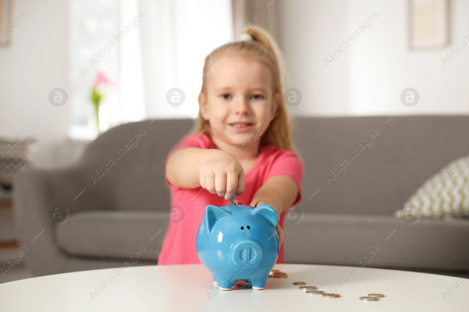 Photo of Cute girl putting coin into piggy bank at table in living room. Saving money
