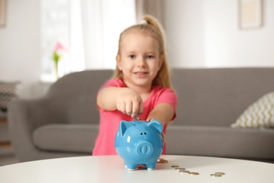 Cute girl putting coin into piggy bank at table in living room. Saving money
