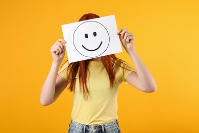 Photo of Woman hiding behind sheet of paper with happy face on yellow background