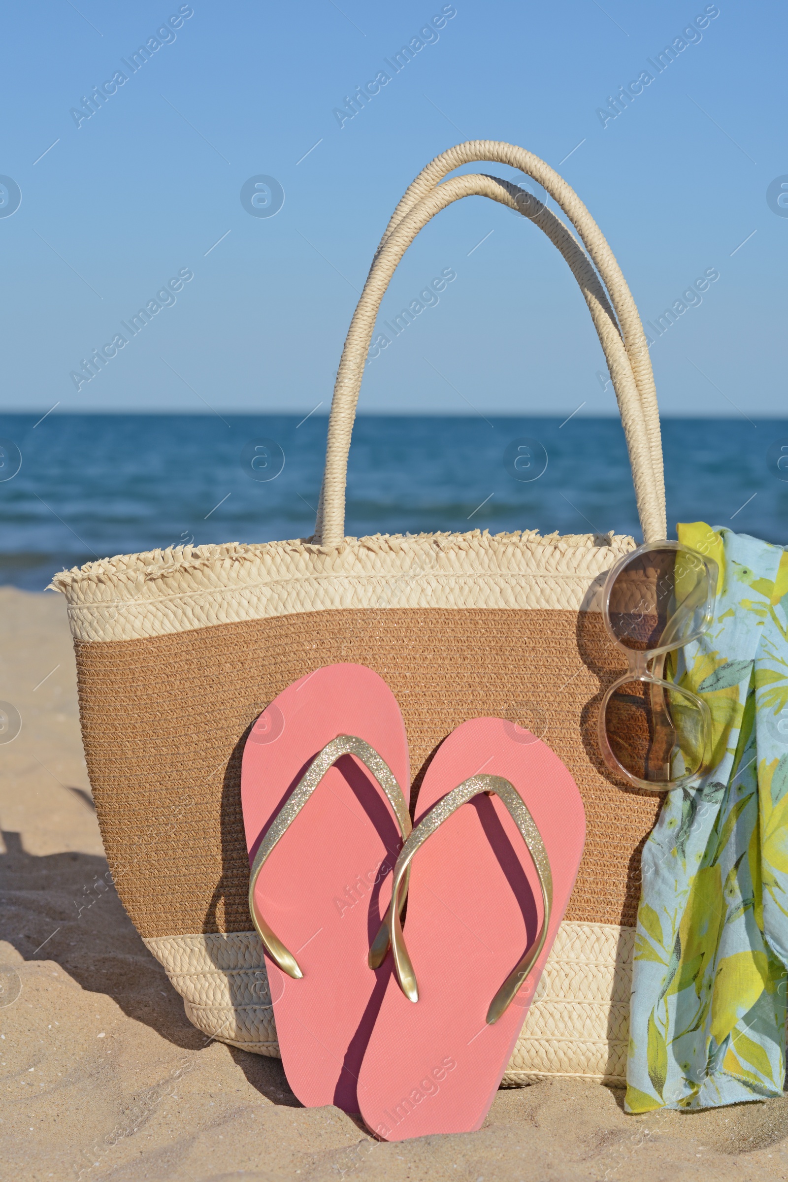Photo of Straw bag with beach wrap, sunglasses and flip flops on sandy seashore. Summer accessories