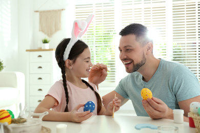 Photo of Happy daughter with bunny ears headband and her father having fun while painting Easter eggs at home