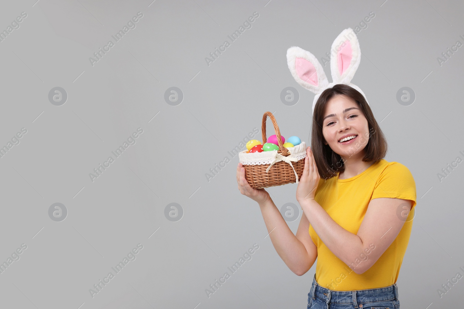 Photo of Easter celebration. Happy woman with bunny ears and wicker basket full of painted eggs on grey background, space for text