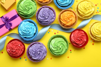 Photo of Delicious birthday cupcakes, gift box, ribbon and sprinkles on yellow background, flat lay