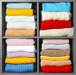 Many knitted winter clothes stacked on shelves near white background