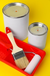 Cans of orange paint, brush, roller and container on yellow background