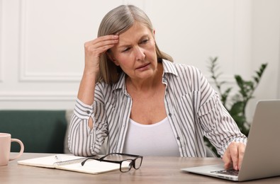 Photo of Menopause. Woman suffering from headache while using laptop at wooden table indoors