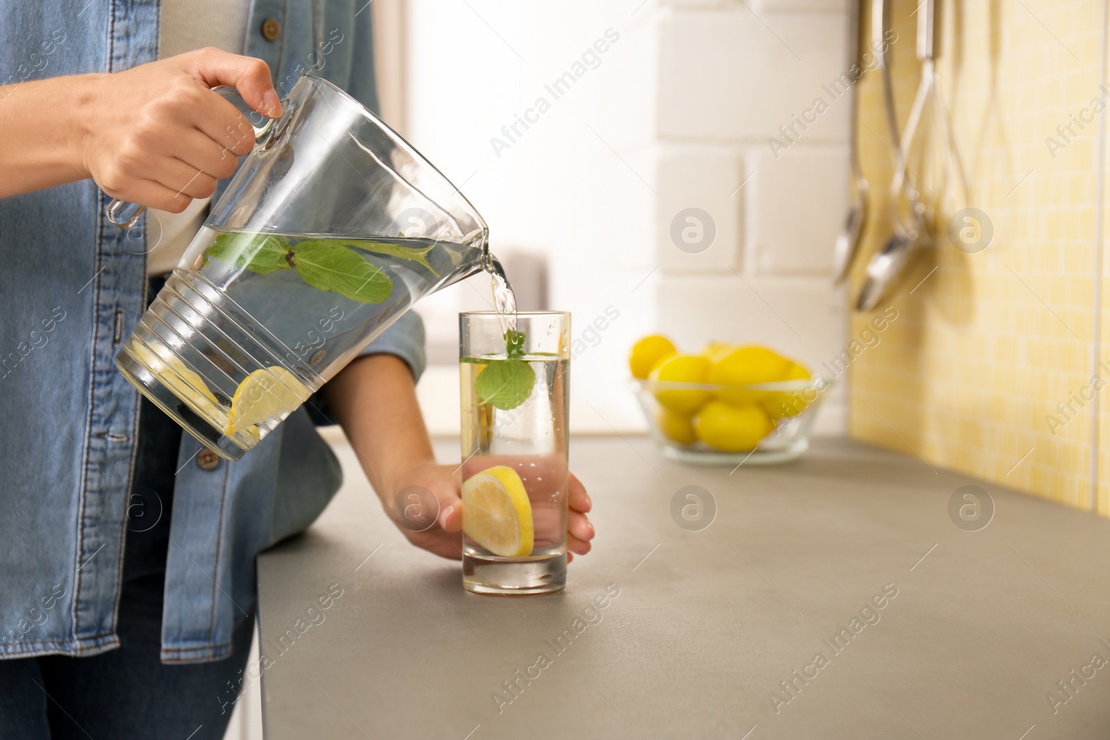 Photo of Young woman pouring lemon water into glass in kitchen, closeup. Space for text