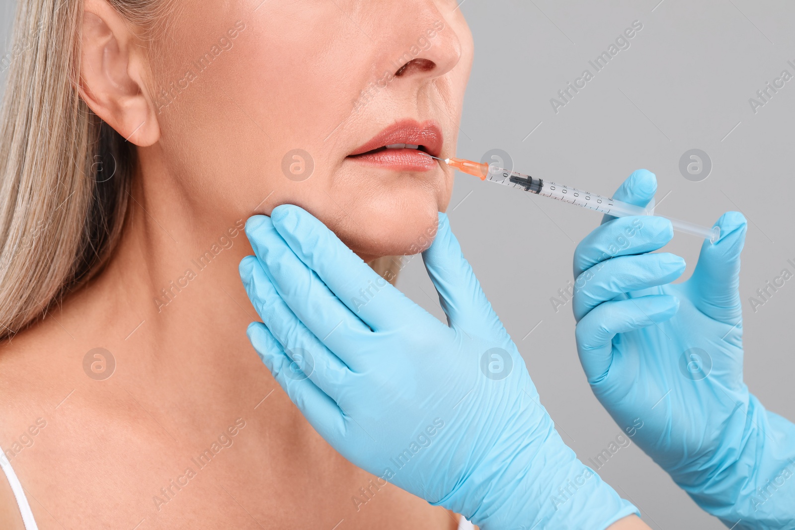 Photo of Doctor giving lips injection to senior woman on light grey background, closeup. Cosmetic surgery