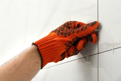 Photo of Man putting plastic cross into joint between ceramic tiles on wall, closeup. Building and renovation works