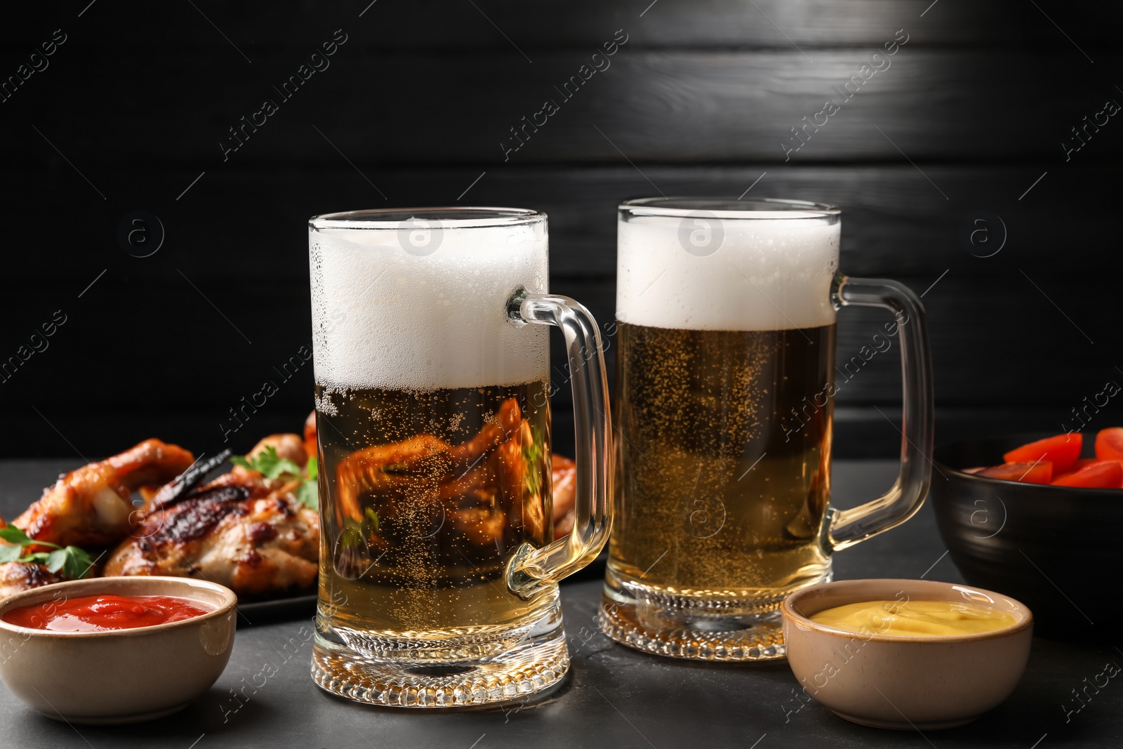 Photo of Mugs with beer, delicious baked chicken wings and sauces on dark grey table