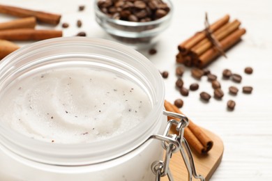 Body scrub in glass jar, cinnamon and coffee beans on white table, closeup
