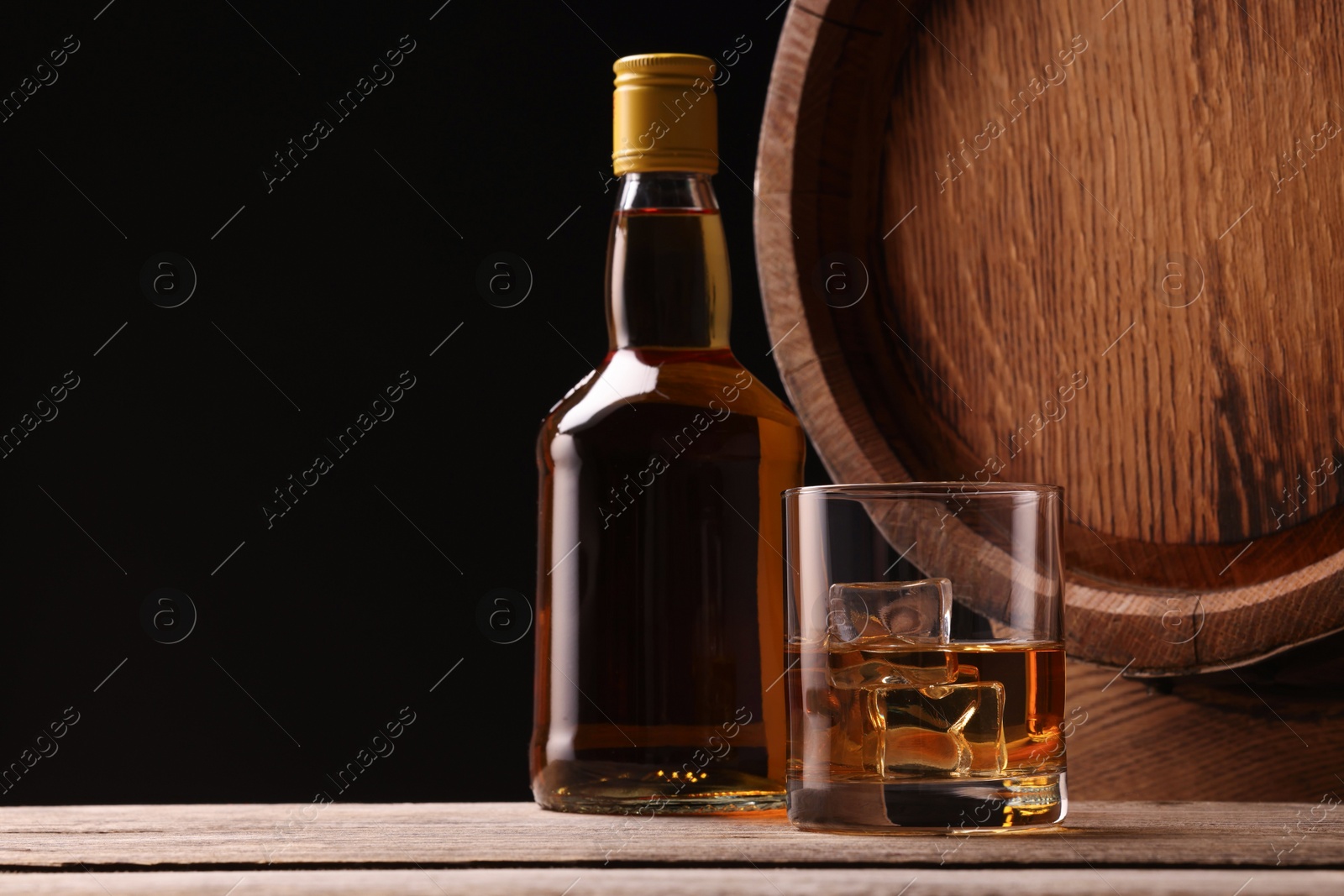 Photo of Whiskey with ice cubes in glass, bottle and barrel on wooden table against black background, space for text