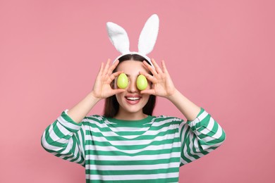 Photo of Happy woman in bunny ears headband holding painted Easter eggs near her eyes on pink background