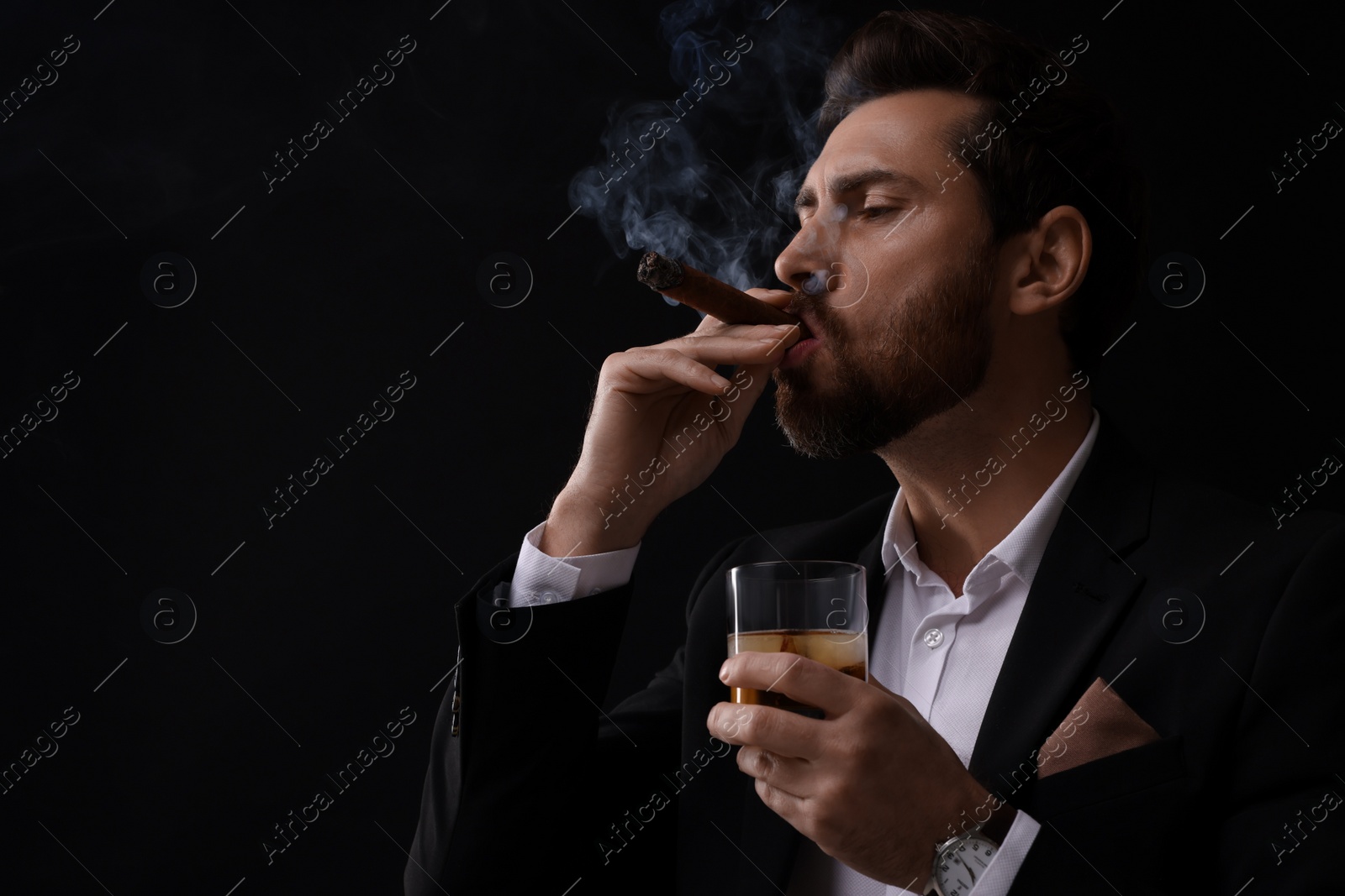 Photo of Handsome man in elegant suit with glass of whiskey smoking cigar on black background. Space for text