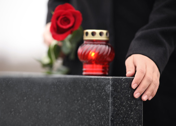 Woman holding red rose near black granite tombstone with candle outdoors, closeup. Funeral ceremony