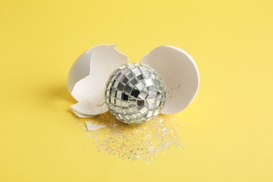 Photo of Silver disco ball, broken eggshell and glitter sprinkles on yellow background