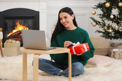 Photo of Celebrating Christmas online with exchanged by mail presents. Smiling woman opening gift box during video call on laptop at home