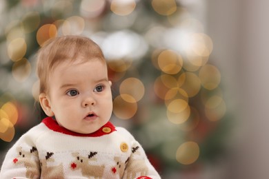 Photo of Cute little baby in Christmas sweater against blurred festive lights, space for text. Winter holiday