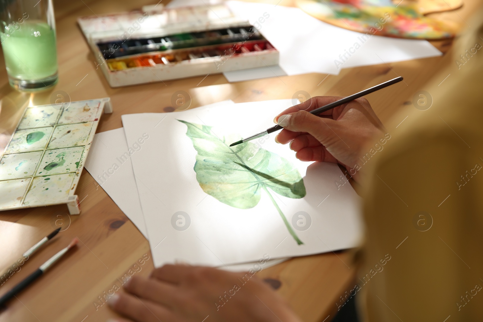 Photo of Young woman drawing leaf with watercolors at table, closeup