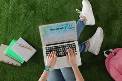 Photo of Woman holding laptop with open travel blogger site on artificial grass, top view