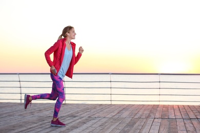 Young woman listening to music and running on pier in morning