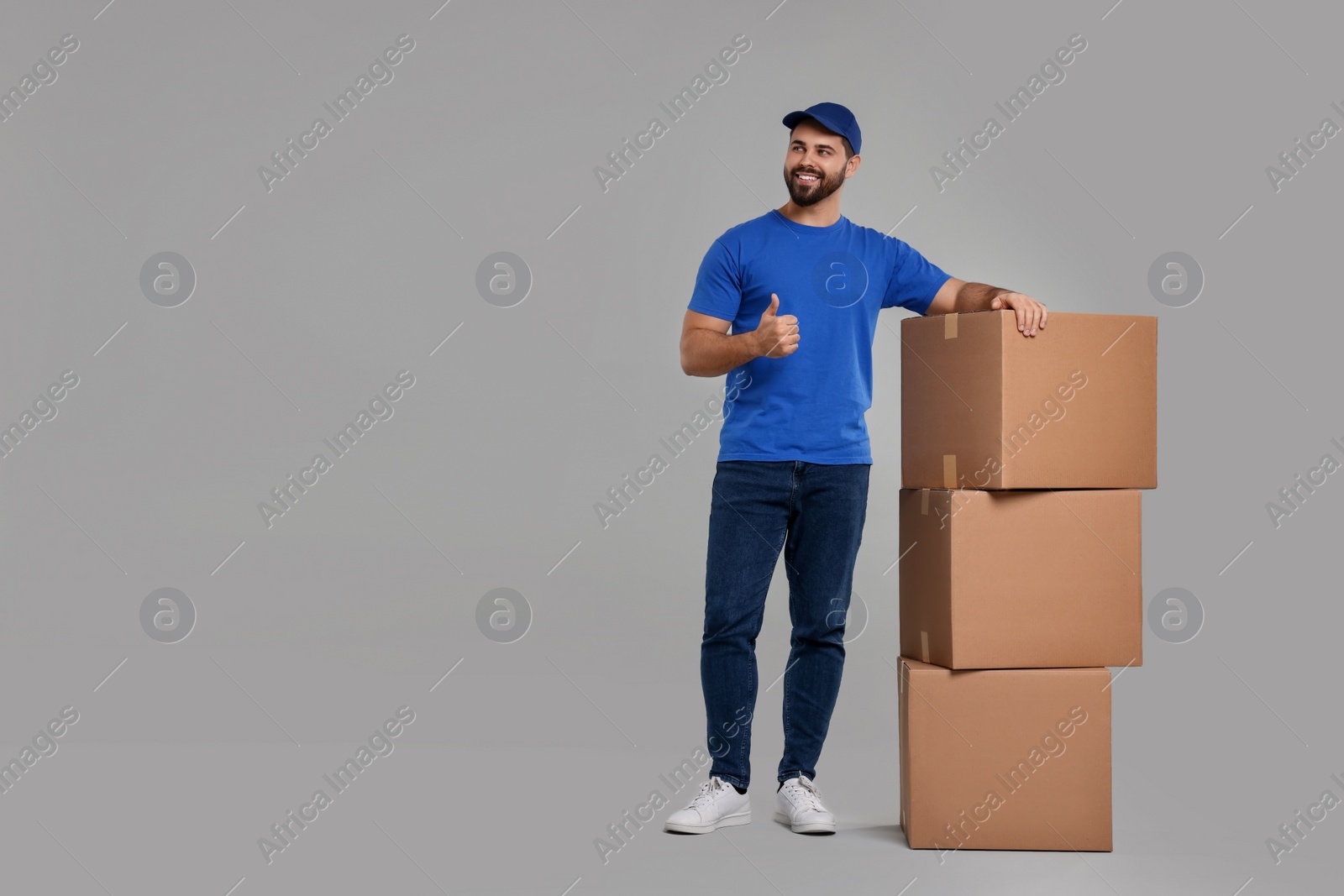 Photo of Happy young courier with stack of parcels showing thumb up on grey background, space for text