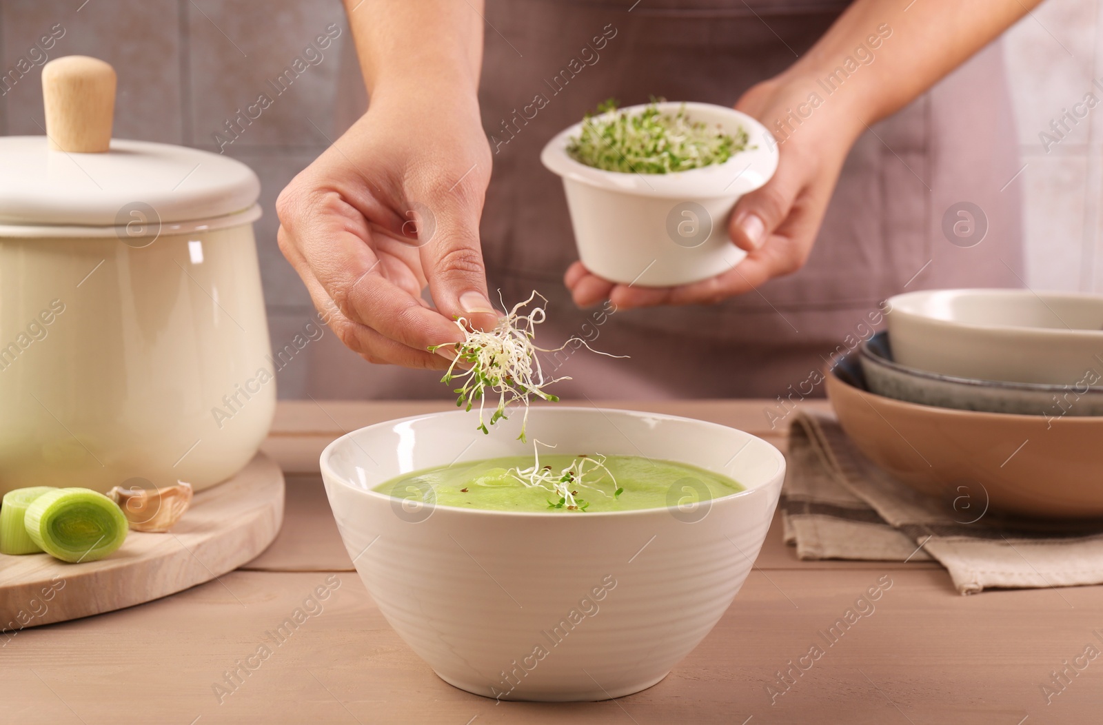 Photo of Woman adding fresh microgreens into bowl with tasty leek soup at wooden table, closeup