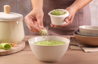 Photo of Woman adding fresh microgreens into bowl with tasty leek soup at wooden table, closeup