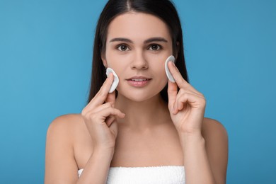 Young woman cleaning face with cotton pads on light blue background