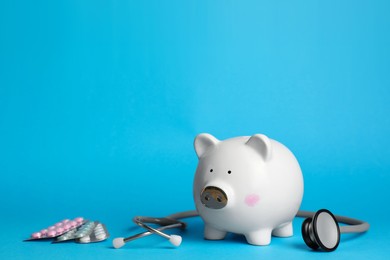 Photo of White ceramic piggy bank, stethoscope and pills on light blue background, space for text. Medical insurance