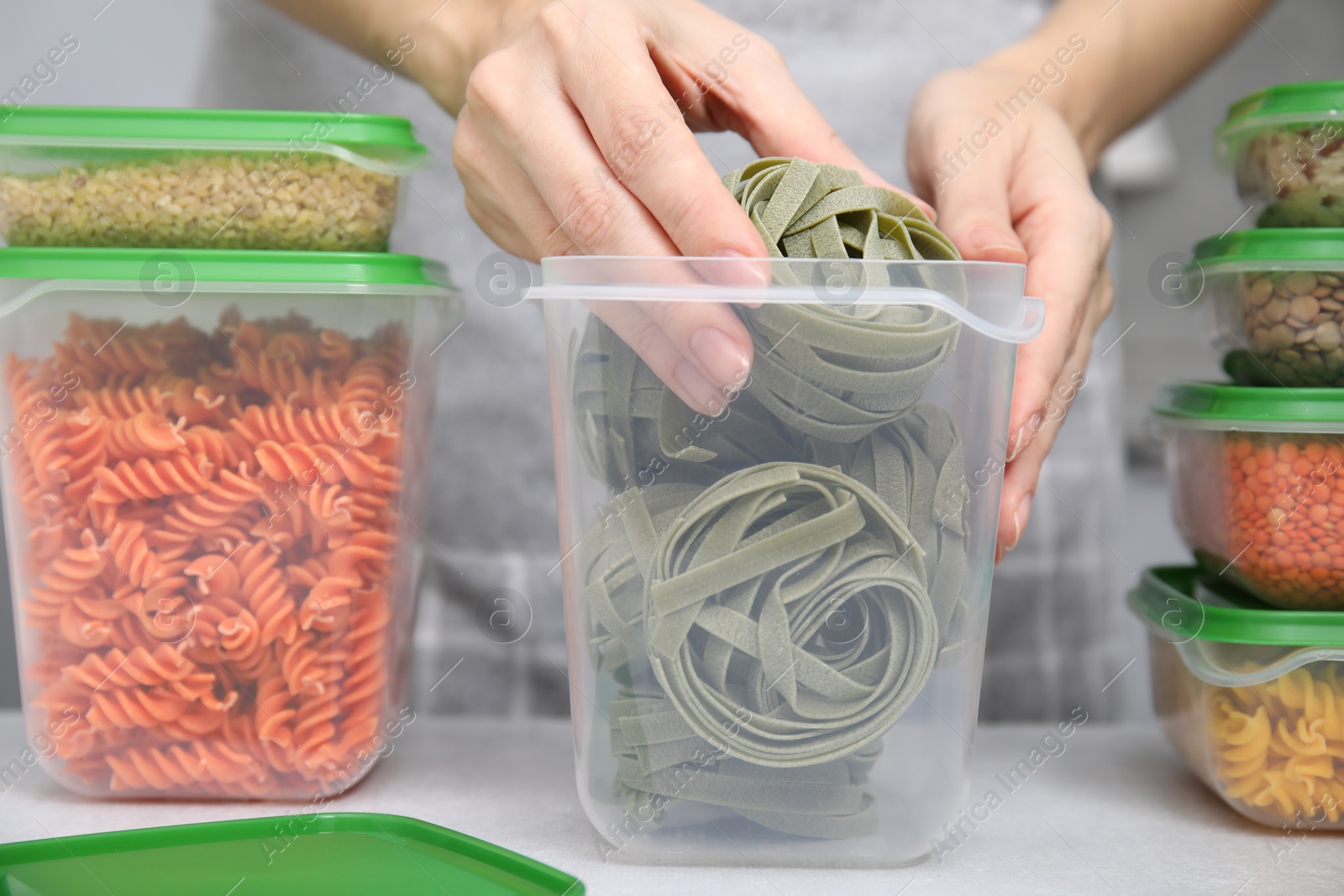 Photo of Woman putting raw pasta into plastic container at light table in kitchen, closeup. Food storage