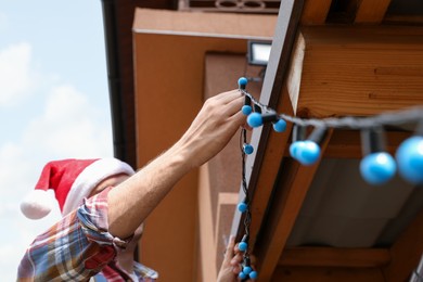 Photo of Man in Santa hat decorating house with Christmas lights outdoors, low angle view