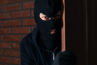 Man in mask spying behind door indoors. Criminal activity