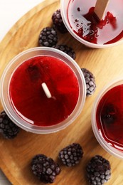 Photo of Tasty berry ice pops in plastic cups on wooden board, flat lay. Fruit popsicle