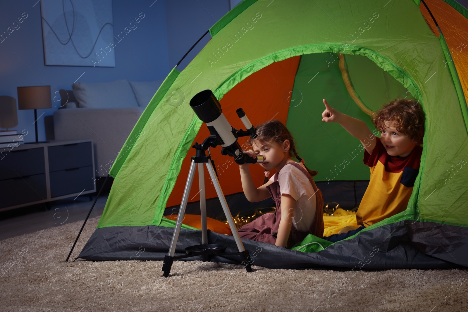 Photo of Cute little children looking at stars through telescope while sitting in camping tent indoors