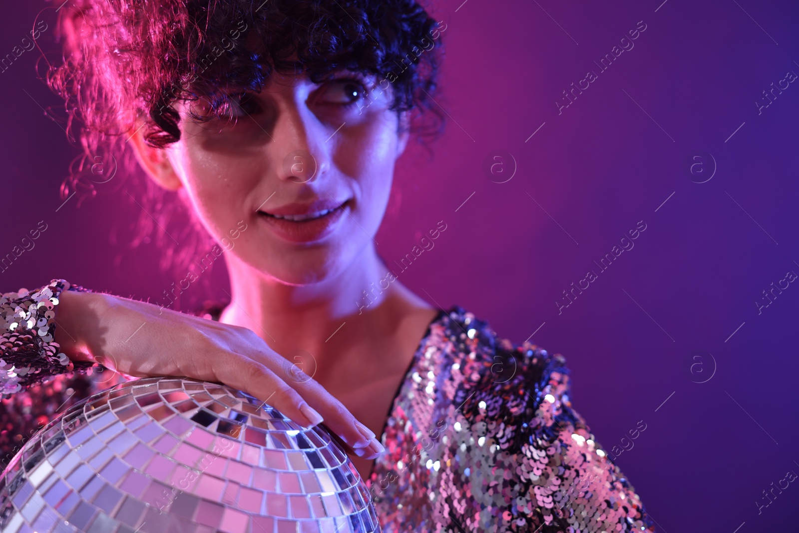 Photo of Beautiful young woman with disco ball posing on color background in neon lights, closeup. Space for text