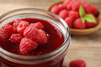 Photo of Glass jar of sweet jam with ripe raspberries on wooden table, closeup