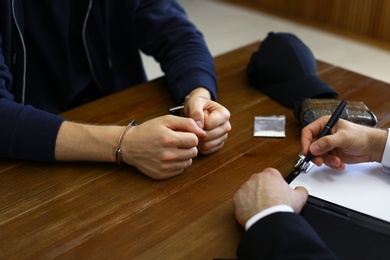 Police officer interrogating criminal in handcuffs at desk indoors