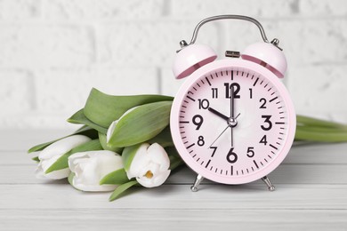 Pink alarm clock and beautiful tulips on white wooden table against brick wall. Spring time