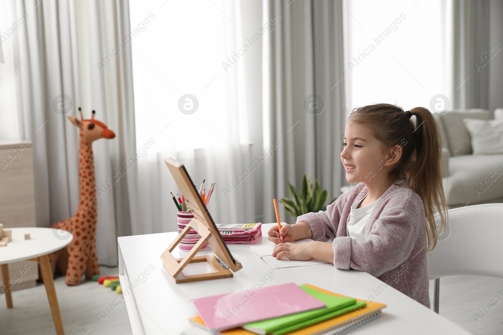 Photo of Adorable little girl doing homework with tablet at table indoors