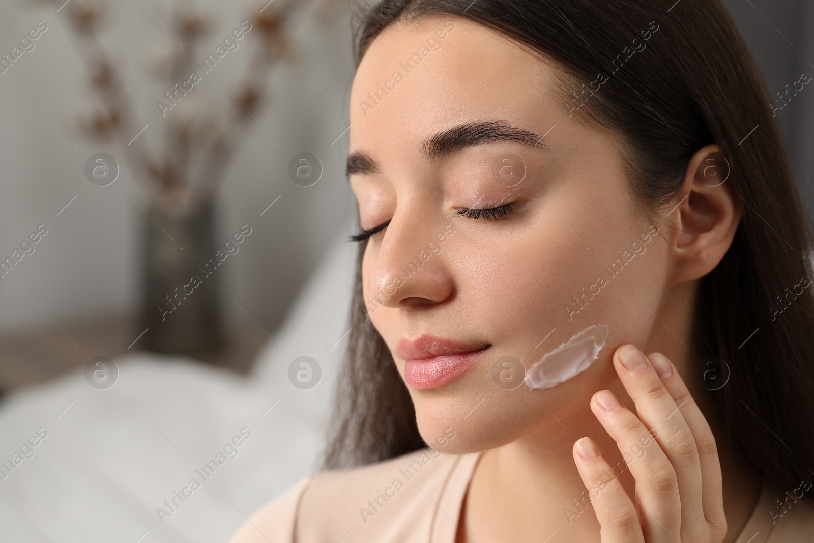 Photo of Young woman with dry skin applying cream onto her face indoors, closeup. Space for text