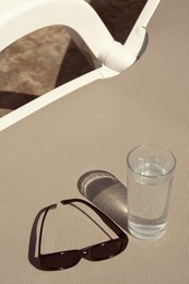 Stylish sunglasses and glass of water on grey sunbed outdoors
