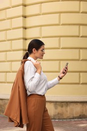 Photo of Young beautiful woman in stylish suit using smartphone near pale yellow wall outdoors