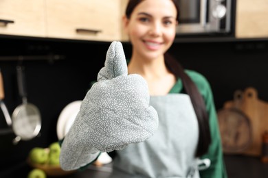Woman in apron and oven glove showing thumb up indoors, focus on hand. Space for text